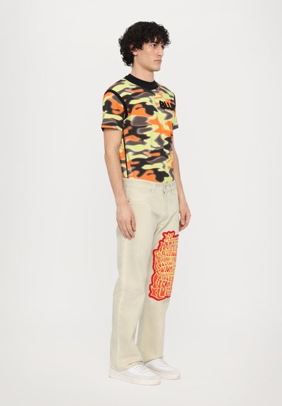 Short-sleeve camouflage shirt in orange, green, and black; paired with light beige pants featuring a large red graphic on the left leg.