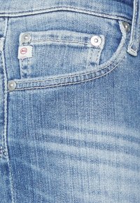 Vaaleansiniset farkut, joissa on etutasku hopeisella metalliosalla, hienovarainen kulutusilme ja AG-logo-tunnus lähellä taskun saumaa.