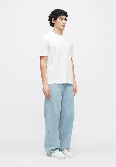 Λευκό κοντομάνικο t-shirt με υφή, συνδυασμένο με ανοιχτό μπλε φαρδιά τζιν και λευκά αθλητικά παπούτσια, μοντέλο στέκεται μπροστά σε απλό φόντο.