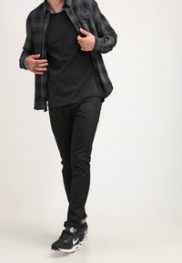 Mężczyzna idący w czarnych trampkach, czarnych wąskich spodniach, czarnym t-shircie i rozpiętej szarej koszuli w kratę, z widocznymi tatuażami na rękach.