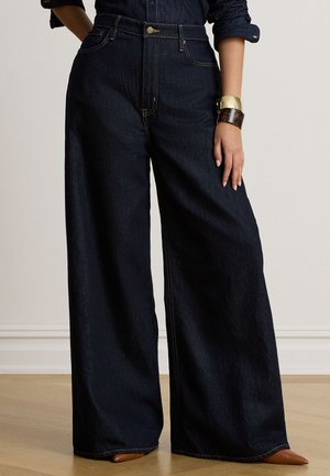 Modèle portant un jean indigo foncé taille haute et à jambes larges, une chemise en denim et des bracelets manchettes dorés.