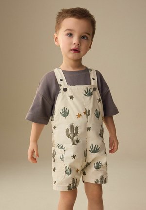 Lille dreng iført beige overalls med kaktus- og stjernemønstre over en løs, mørkegrå t-shirt, stående mod en ensfarvet beige baggrund.