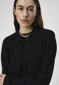 Sort cardigan med krave, der har knapper foran og ribbet tekstur. Sammen med en sort ribbet top og en sølvkæde.