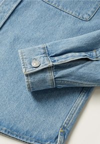 Manche de chemise en denim bleu clair avec un poignet boutonné reposant sur un fond en tissu denim assorti.