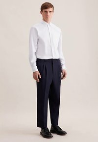 Camicia bianca a maniche lunghe con design a bottoni, abbinata a pantaloni plissettati blu navy scuro e scarpe eleganti nere. Stile semplice e pulito.