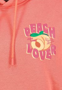 Pfirsichfarbener Hoodie mit einem Grafikdesign, auf dem der Text "PEACH LOVER" und eine Pfirsichillustration zu sehen sind. Das Material wirkt weich mit einer strukturierten Oberfläche.
