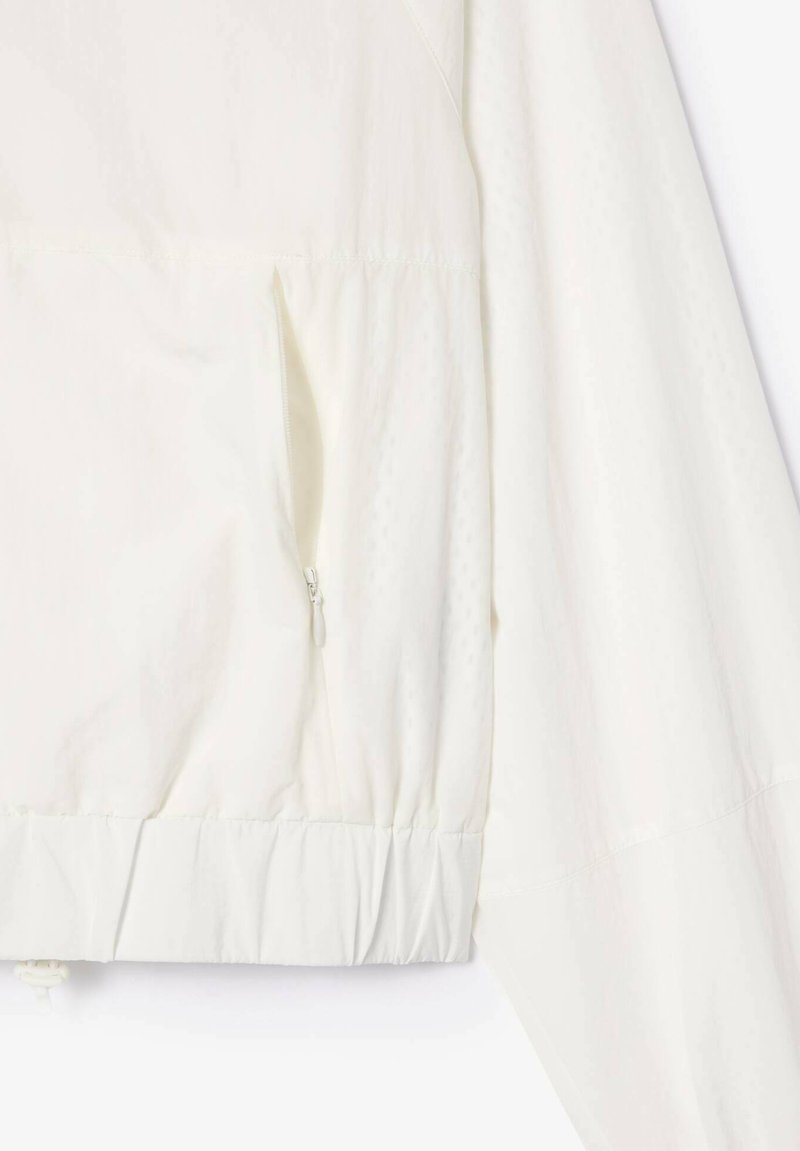 Zbliżenie na rękaw białej kurtki i bok, z elastycznym wykończeniem i kieszenią na zamek błyskawiczny.