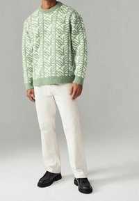 Pull vert clair à motifs géométriques, col rond, poignets côtelés, associé à un pantalon crème et des chaussures noires épaisses.