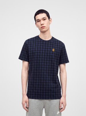 Jonge man draagt een donker patroon T-shirt met een klein kleurrijk logo op de borst en lichtgrijze Nike joggingbroek, staat neutraal.