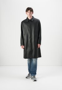 Schwarze Lederjacke mit Kragen, langen Ärmeln und Vordertaschen; hat einen geraden Schnitt und kontrastierende Nähte. Getragen über blauen Jeans und Turnschuhen.