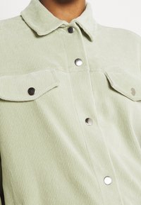 Giacca in corduroy verde chiaro con colletto, bottoni a pressione argentati e due tasche sul petto. Presenta una texture a righe verticali.