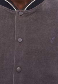 Veste en tissu côtelé gris foncé avec des boutons-pression et un petit logo colibri brodé près de la poitrine, col rayé visible.