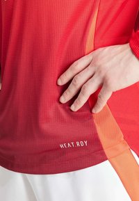 Haut de sport rouge avec tissu texturé, présentant des accents orange. Logo "HEAT.RDY" en blanc en bas à droite. Vue rapprochée avec la main placée.