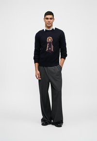 Mornarica pulover s pletenim dizajnom psa i "H" logom, uparen s sivim hlačama širokih nogavica i crnim cipelama. Cjelokupna glatka tekstura.