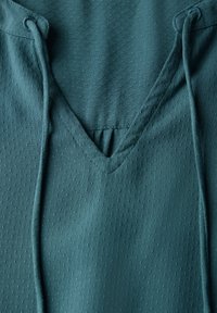 Teal atletisk top lavet af struktureret stof med v-hals og snore. Har små perforeringer for åndbarhed.