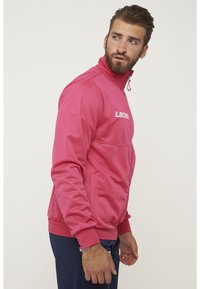 Rosa zip-up jacka med ståkrage och vit logotyp, med en framficka och ribbad mudd och nederkant. Slät textur och atletisk passform.