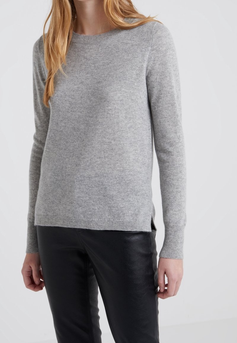 Femme portant un pull gris clair à manches longues et un pantalon en cuir noir, debout devant un fond blanc uni. Son visage n'est pas visible.