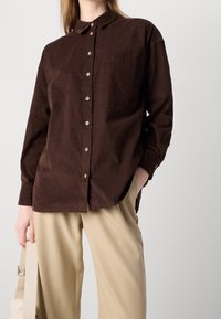 Camicia button-up in corduroy marrone con colletto, dotata di una tasca frontale e pantaloni beige a gamba larga. Tessuto texturizzato e vestibilità rilassata.