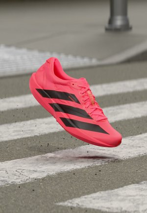 Zapatilla de correr rosa brillante con rayas negras en el aire sobre un paso de peatones en pavimento de asfalto.