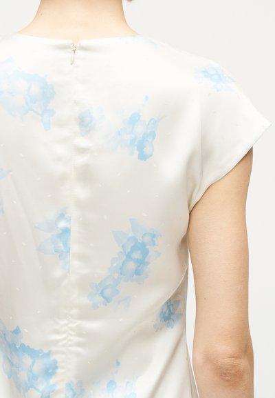 Πλάτη ατόμου που φοράει λευκή μπλούζα με ανοιχτό μπλε λουλουδάτο μοτίβο και κοντά μανίκια με θυρεό, πάνω σε απλό φόντο.