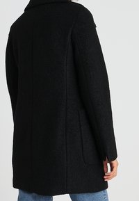 Svart ullkappa med en strukturerad design, som har en klädd krage, långa ärmar och sidofickor, texturerad finish och medellång längd.