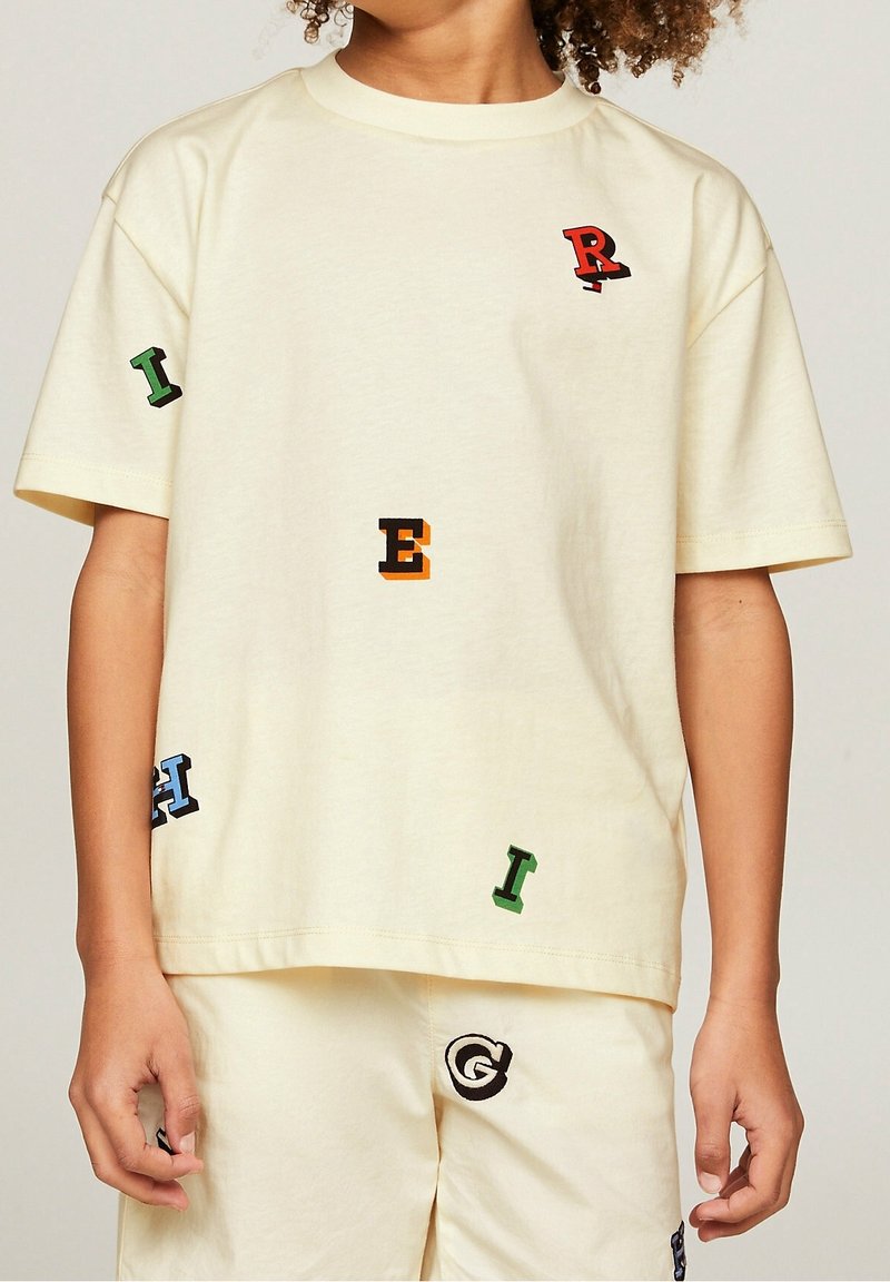Cremfarvet bomulds T-shirt med korte ærmer, prydet med farverige broderede bogstaver (R, E, I, H) spredt over fronten og siderne.