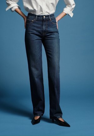 Person trägt dunkelblaue Jeans mit hohem Bund, weißes langärmliges Hemd mit hochgekrempelten Ärmeln und schwarze spitze High Heels, steht mit den Händen in den Hüften.