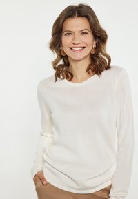 Leichter, langärmeliger Creme-Pullover mit rundem Ausschnitt und gerippter Textur. Kombiniert mit beigefarbenen Hosen, die eine lockere Passform bieten.