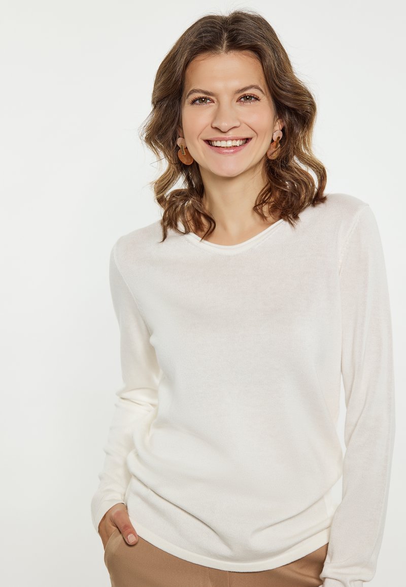 Leichter, langärmeliger Creme-Pullover mit rundem Ausschnitt und gerippter Textur. Kombiniert mit beigefarbenen Hosen, die eine lockere Passform bieten.
