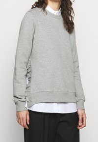 Grauer Sweatshirt mit Rundhalsausschnitt, strukturierter Stoff, geraffte Seitendetails und gerippte Bündchen sowie Saum, über einem weißen Shirt getragen.