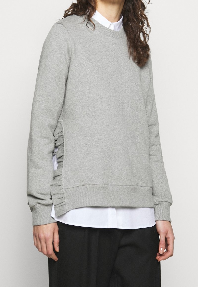 Grauer Sweatshirt mit Rundhalsausschnitt, strukturierter Stoff, geraffte Seitendetails und gerippte Bündchen sowie Saum, über einem weißen Shirt getragen.