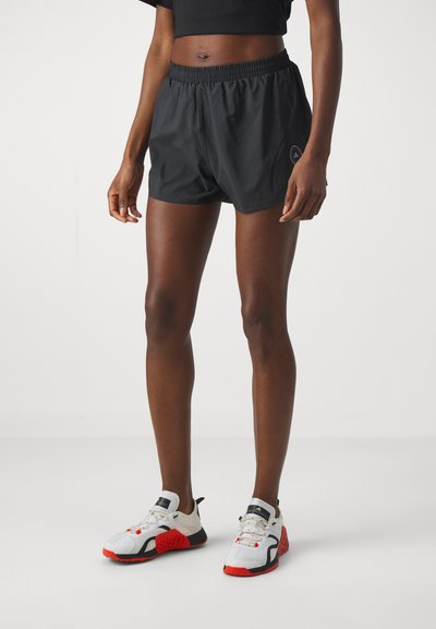 Person trägt schwarze Sportshorts, ein schwarzes kurzes Oberteil und weiße Sneakers mit roten und schwarzen Details und steht vor einem schlichten Hintergrund.