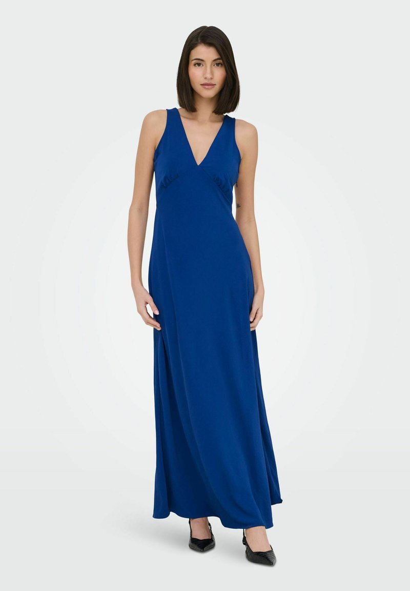 Femme debout portant une robe longue bleu foncé sans manches avec un col en V et des talons noirs pointus sur un fond clair uni.