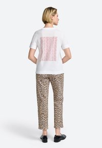 Witte katoen t-shirt met rode tekstafdruk op de achterkant, gecombineerd met lichtbruine broek met luipaardprint en zwarte platte schoenen.