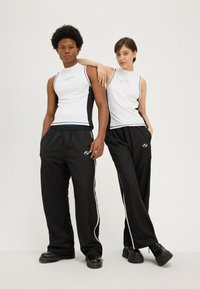 Dwie modelki noszą sportowe stroje: białe bezrękawniki z czarnymi bocznymi panelami, czarne spodnie z białymi paskami oraz masywne czarne obuwie.