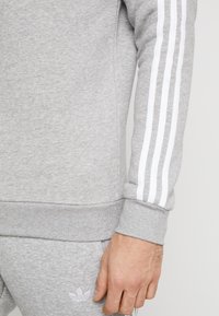 Närbild av en persons arm och hand som bär en grå sweatshirts och sweatpants med vita tredubbla ränder på ärmen och Adidas-logotypen på byxorna.