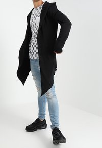 Giacca lunga nera con cappuccio, camicia grigia a scacchi, jeans chiari strappati blu e sneakers nere. Presenta maniche lunghe ed orlo asimmetrico.
