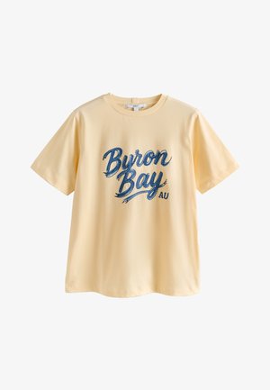 Maglietta giallo chiaro a maniche corte con la scritta "Byron Bay AU" in grande testo corsivo blu davanti.