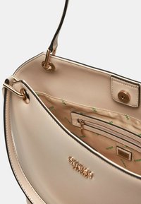 Interior de bolso de mano de imitación cuero color crema con herrajes dorados, bolsillo interior con cremallera y forro de tela de marca que presenta Logos verdes de "GUESS".