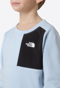 Sudadera azul claro con un panel negro que presenta el logo de The North Face en blanco. Textura suave, cuello redondo y detalle de costuras en contraste.