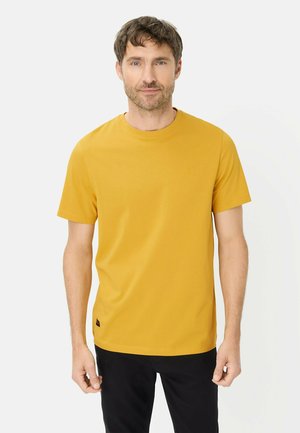 Mann mit kurzen braunen Haaren, der ein senfgelbes kurzärmeliges T-Shirt und schwarze Hosen trägt, steht vor einem schlichten hellen Hintergrund.