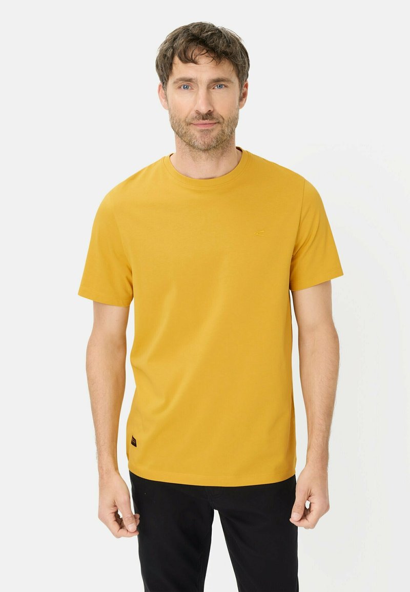 Mann mit kurzen braunen Haaren, der ein senfgelbes kurzärmeliges T-Shirt und schwarze Hosen trägt, steht vor einem schlichten hellen Hintergrund.