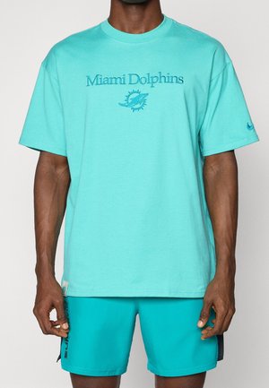 Mand iført en turkis Miami Dolphins t-shirt og matchende shorts står foran en ensfarvet baggrund.