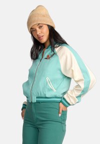 Teal and cream bomber jacket with striped cuffs, zipper front, and embroidered detail. Pairing with matching teal pants and beige ribbed beanie.