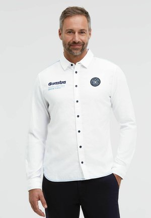 Man with short hair and beard wearing a white button-up shirt with logos, standing with one hand in pocket against plain background.