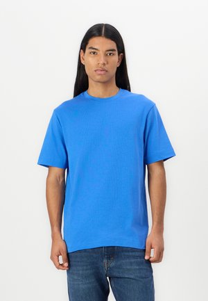 T-shirt en coton bleu à manches courtes, col rond et coupe décontractée. Associé à un jean en denim, le tout sur un fond blanc uni.