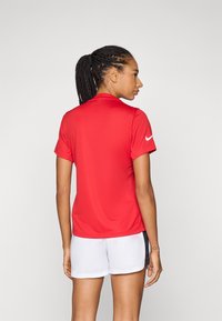 Rød athletic polo-skjorte med kortermet, glatt tekstur og krage. Har en hvit Nike-logo på venstre erme. Bæres med hvite shorts.