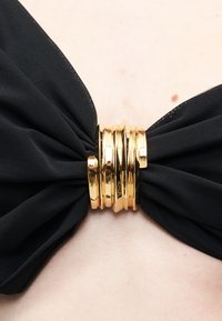 Μαύρο ύφασμα μαζεμένο και συγκρατημένο από πολλούς γυαλιστερούς χρυσούς κρίκους πάνω σε ανοιχτόχρωμο δέρμα με μικρές φακίδες ορατές.