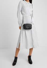 Musta olkalaukku, jossa on kultainen logo, suorakulmainen muoto, teksturoitu nahka ja ketjuolkaimet. Yhdistetty valkoiseen raidalliseen mekkoon ja nilkkureihin.