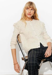 Gilet en maille crème avec des motifs complexes, col rond, fermeture à boutons et manches volantes. Le modèle l'a associé à un pantalon à carreaux.